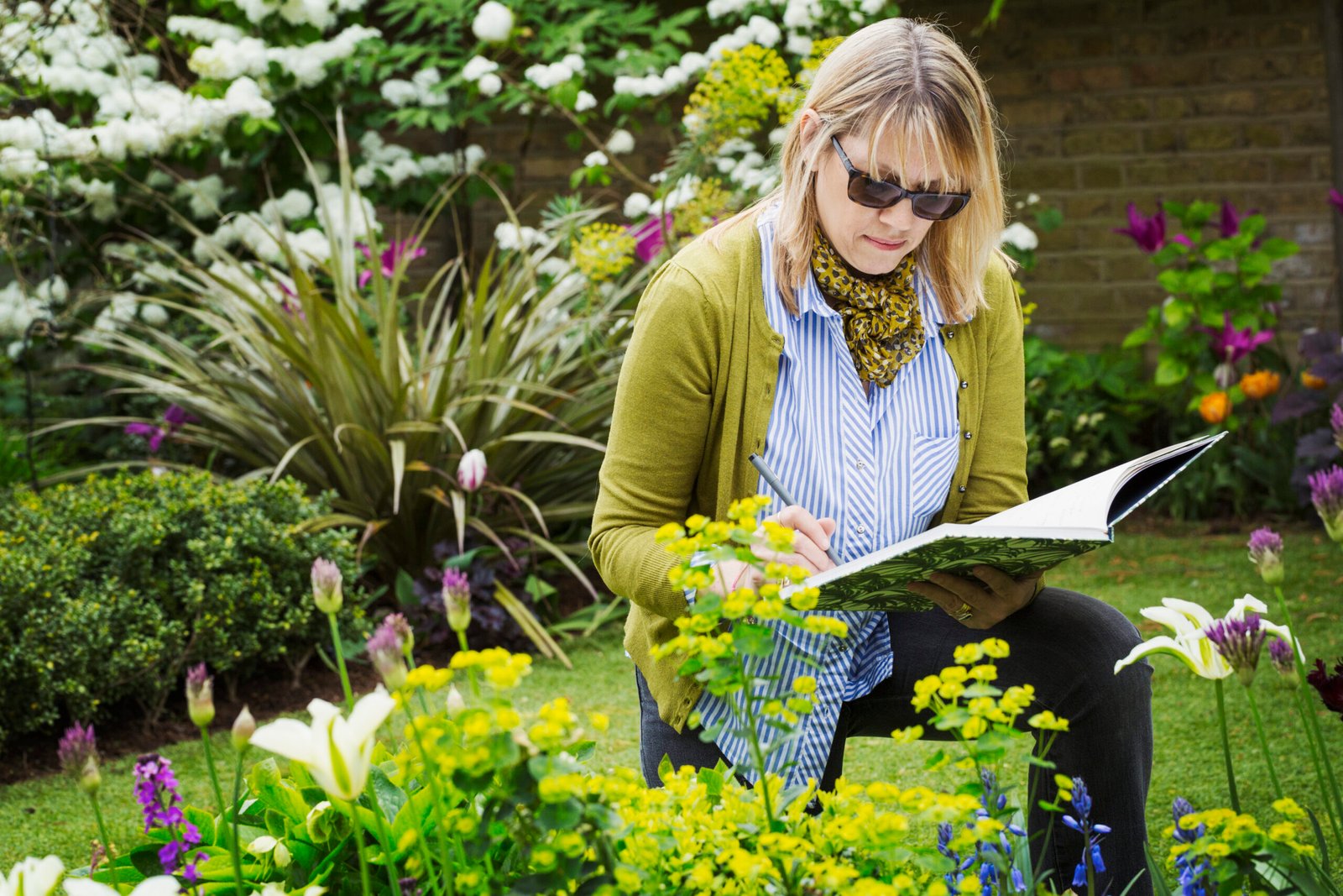 Woman wearing sunglasses standing in a garden by a flowerbed, drawing in a sketchbook.,Garden Design,UK